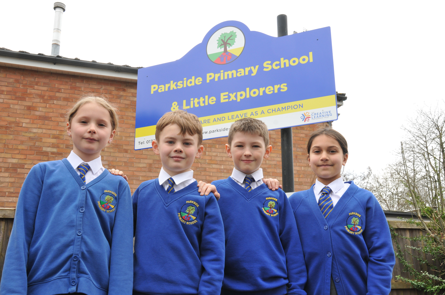Four elementary students stand in front of a Parkside Primary School sign. They wear royal blue uniforms with the school emblem. Sign reads: "Parkside Primary School & Little Explorers, DARE AND LEAVE AS A CHAMPION".