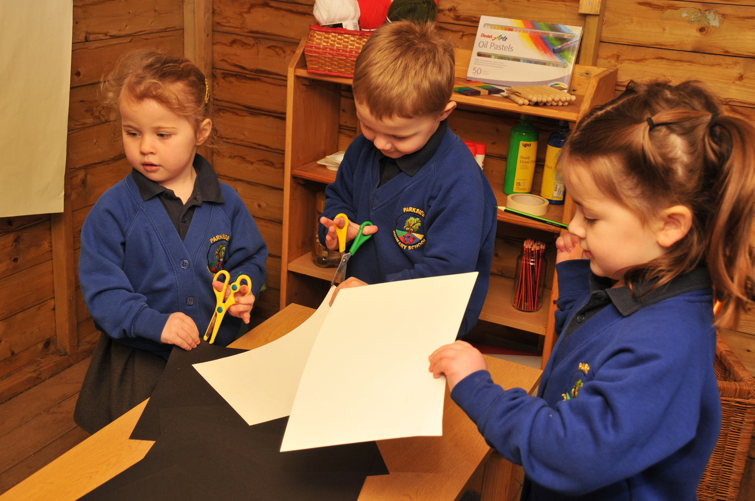 Three Parkside children in school uniforms cut paper at a table. Art supplies and a basket are on a shelf behind them.