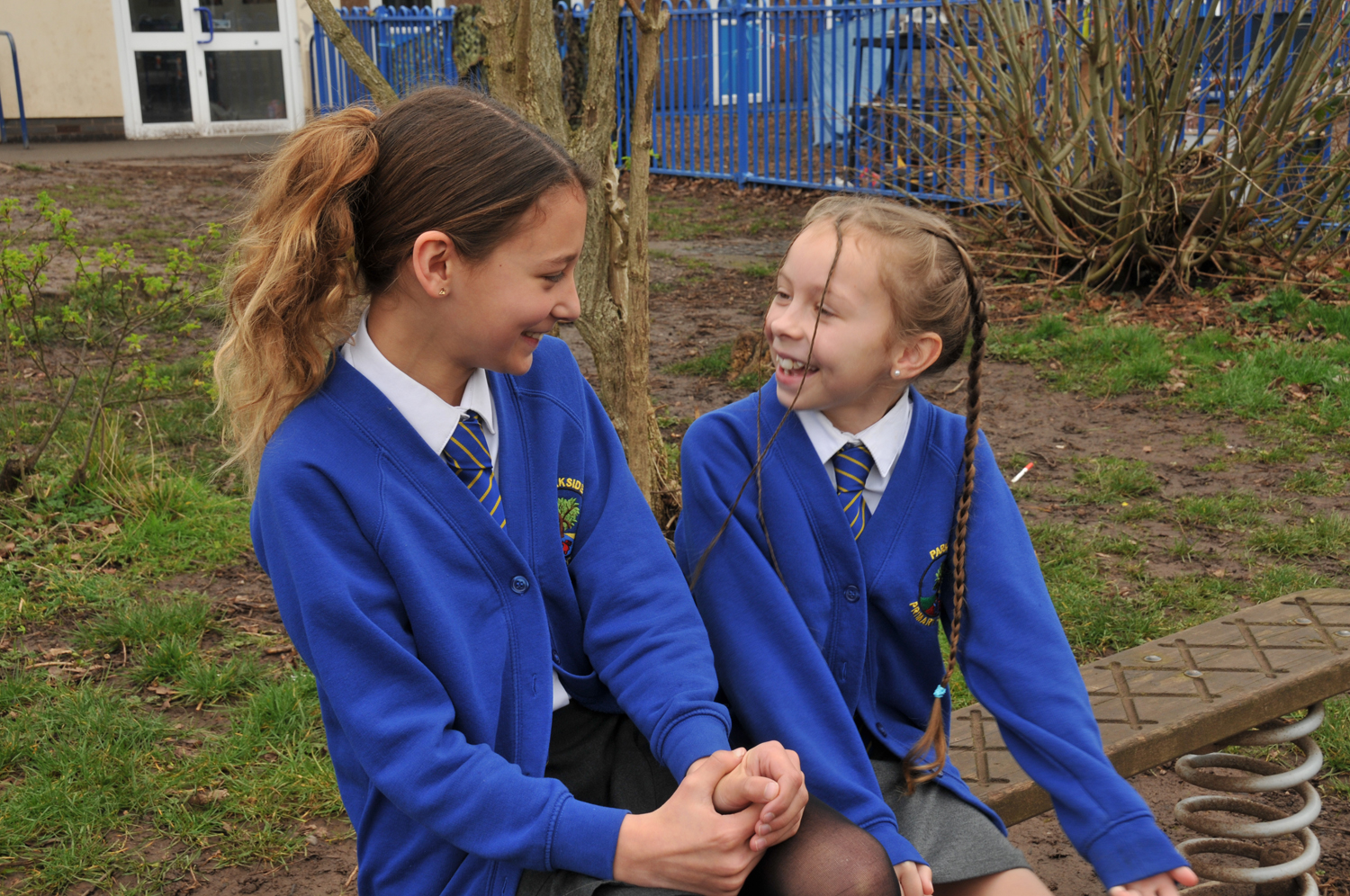 Two students in Parkside Primary uniforms smile at each other. They sit outside on the playground, enjoying a cheerful moment.