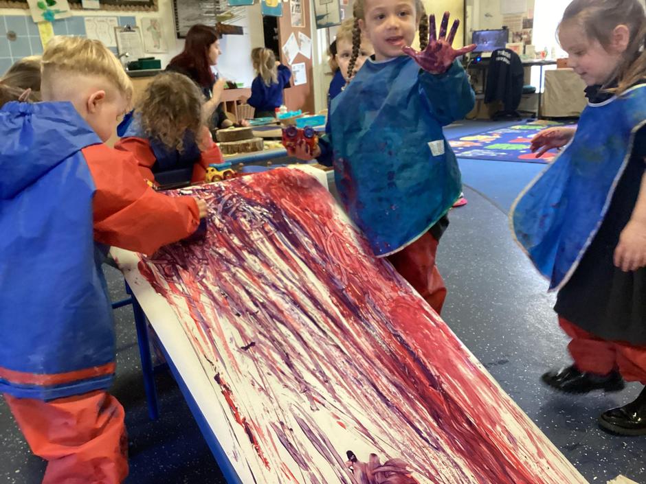 Children at the Little Explorers Nursery wear aprons while finger painting on a large easel. Red and purple paint streaks across the paper as a child holds their painted hand in the air.