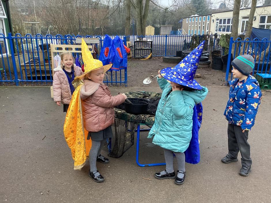 Four young children wearing hats and capes, stand around a small table in an outdoor play area, pretending to cast spells into a pot.